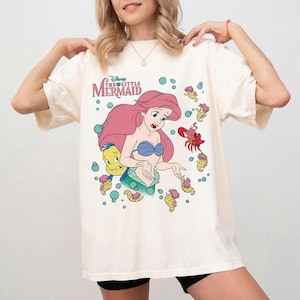 May include: An off-white short-sleeve t-shirt featuring a graphic print of Ariel from The Little Mermaid, along with Flounder, Sebastian, and several seahorses. The design includes teal bubbles and the text Disney The Little Mermaid in red.
