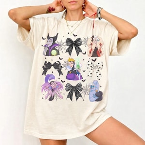 May include: Cream-colored t-shirt featuring cartoon illustrations of Disney villains. The design includes characters like Maleficent, the Evil Queen, and Ursula, along with black bows, spiderwebs, and bats. The shirt has a relaxed fit.