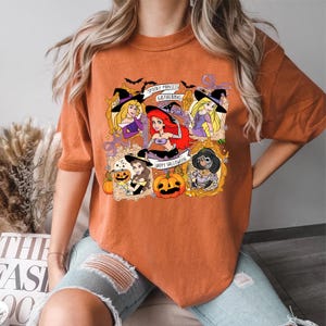 May include: Orange t-shirt featuring a Halloween-themed graphic with cartoon princesses in witch hats and pumpkins. The text on the shirt reads "Spooky Princess Gathering" and "Happy Halloween".