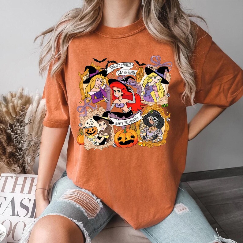May include: Orange t-shirt featuring a Halloween-themed graphic with cartoon princesses in witch hats and pumpkins. The text on the shirt reads "Spooky Princess Gathering" and "Happy Halloween".