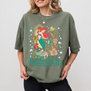 Op de afbeelding: Olijfgroen T-shirt met een afbeelding van Ariel uit De Kleine Zeemeermin. Het ontwerp toont Ariel met rood haar en een groene staart, en de tekst "THE LITTLE MERMAID" in turquoise. Het T-shirt heeft een losse pasvorm.