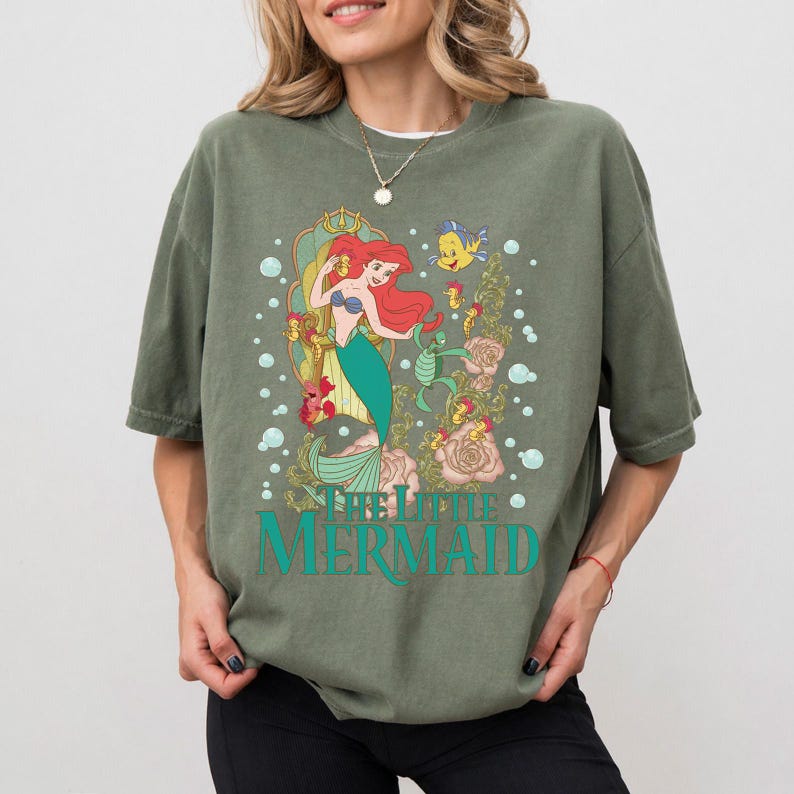 Op de afbeelding: Olijfgroen T-shirt met een afbeelding van Ariel uit De Kleine Zeemeermin. Het ontwerp toont Ariel met rood haar en een groene staart, en de tekst "THE LITTLE MERMAID" in turquoise. Het T-shirt heeft een losse pasvorm.