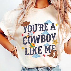 May include: A cream-colored t-shirt featuring the dark blue text "YOU'RE A COWBOY LIKE ME" surrounded by colorful illustrations of a brown cowboy hat, a rocket ship, a yellow ball, a yellow sheriff star badge, brown cowboy boots, and stars.