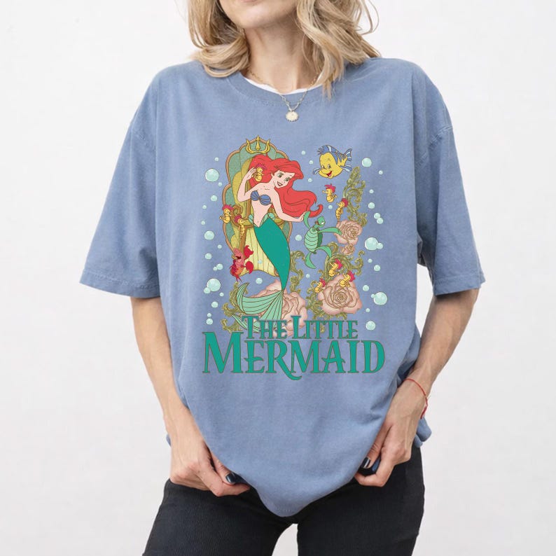 Op de afbeelding: Lichtblauw T-shirt met een kleurrijke afbeelding van Ariel uit De Kleine Zeemeermin. Het ontwerp toont Ariel met rood haar en een groene staart, omringd door bloemen en de tekst "The Little Mermaid" in groen.