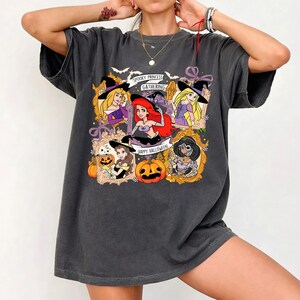May include: Dark gray t-shirt featuring a Halloween-themed graphic. The graphic includes cartoon princesses in witch hats, pumpkins, and the text "Spooky Princess Gathering" and "Happy Halloween". The shirt has a relaxed fit.