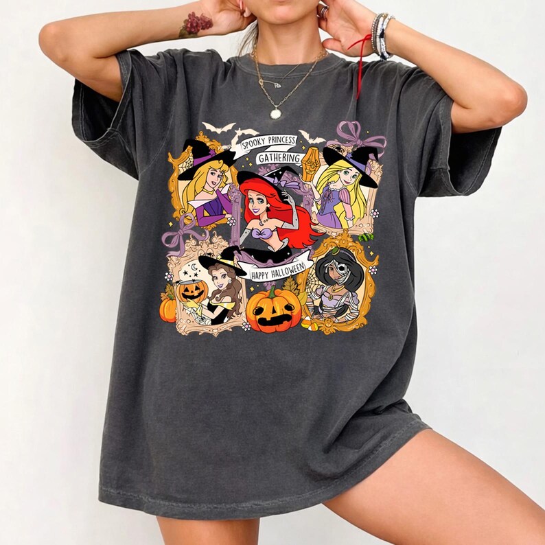 May include: Dark gray t-shirt featuring a Halloween-themed graphic. The graphic includes cartoon princesses in witch hats, pumpkins, and the text "Spooky Princess Gathering" and "Happy Halloween". The shirt has a relaxed fit.