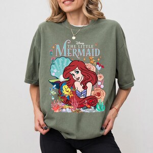 Op de afbeelding: Een olijfgroen T-shirt met een kleurrijke afbeelding van Ariel uit Disney's De Kleine Zeemeermin. Het ontwerp bevat Ariel, Botje en diverse zee-elementen. De tekst "Disney The Little Mermaid" staat boven de afbeelding.