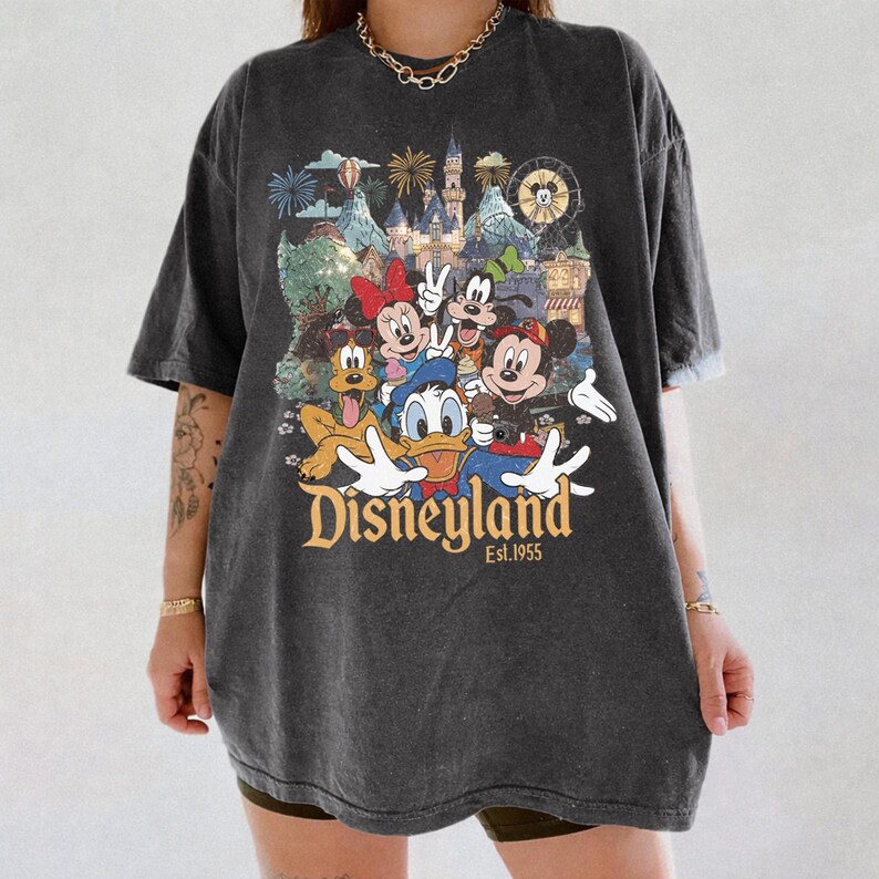 May include: A charcoal gray short-sleeve t-shirt featuring a colorful graphic print of Mickey Mouse, Minnie Mouse, Donald Duck, Goofy, and Pluto in front of a castle. The text Disneyland Est. 1955 is printed in gold below the characters.