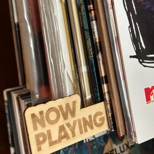 May include: A wooden "NOW PLAYING" record divider sits among a collection of vinyl records. The divider is light brown with the words "NOW PLAYING" etched into the wood. Album covers are visible, including "UNPLUGGED IN NEW YORK" and "DELUXE EDITION."