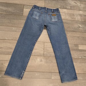 May include: Light blue denim jeans with a classic five-pocket design. The jeans have a straight leg cut and a faded appearance, with visible stitching and brass-colored rivets. The back pockets feature a simple, rectangular design, and a leather patch is visible.