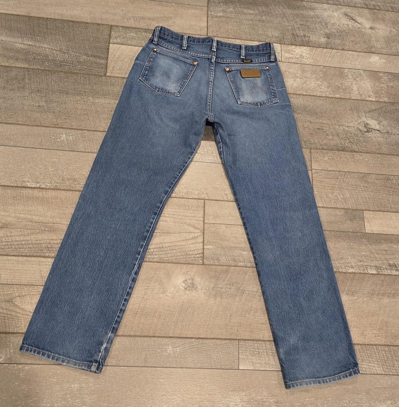 May include: Light blue denim jeans with a classic five-pocket design. The jeans have a straight leg cut and a faded appearance, with visible stitching and brass-colored rivets. The back pockets feature a simple, rectangular design, and a leather patch is visible.
