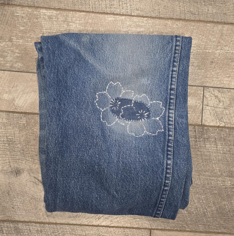 May include: Folded blue denim jeans with a white floral embroidery design on the front. The embroidery features a detailed flower pattern with delicate stitching. The jeans are folded neatly, showcasing the design.