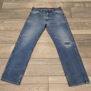 May include: Faded blue denim jeans with a distressed knee. The jeans have a classic five-pocket design, a button and zipper closure, and a straight leg. The denim has a worn appearance.