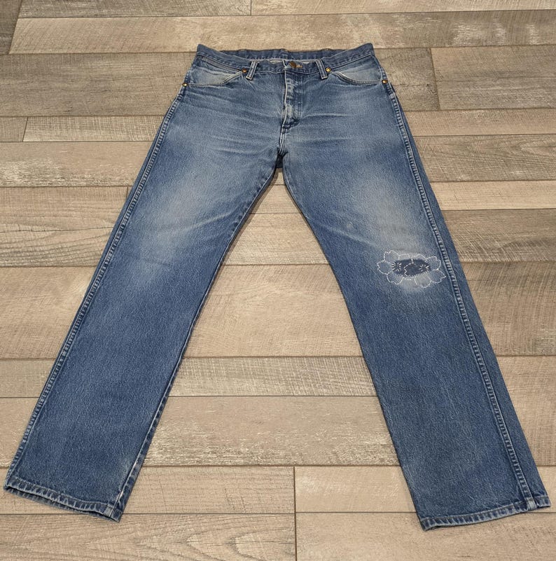 May include: Faded blue denim jeans with a distressed knee. The jeans have a classic five-pocket design, a button and zipper closure, and a straight leg. The denim has a worn appearance.