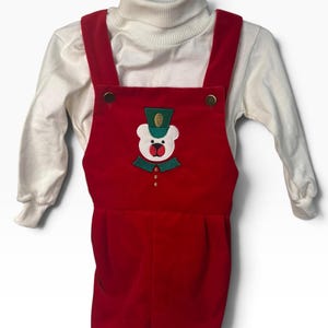 May include: A red overall set featuring a white turtleneck long-sleeve shirt underneath. The red overalls have a central embroidered bear graphic wearing a green hat and collar, with gold-tone buttons at the shoulders and chest.