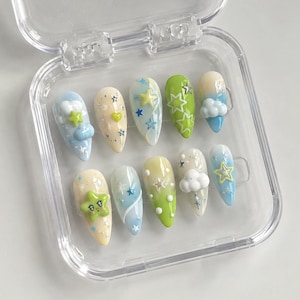 May include: A collection of ten artificial nails in a clear plastic case. Each nail showcases a unique design, incorporating pastel shades, stars, clouds, and other celestial-themed embellishments. The nails are almond-shaped and ready for use.