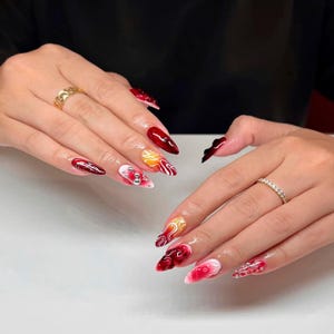 Puede incluir: Uñas acrílicas largas y puntiagudas con varios diseños. Las uñas presentan una mezcla de colores rojo, blanco y amarillo, algunas adornadas con adornos plateados. Una mano tiene un anillo de oro y la otra un anillo de diamantes.