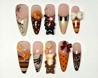 Op de afbeelding: Een set van tien kunstnagels met verschillende ontwerpen. De nagels hebben een nude basis met gedetailleerde kunst, waaronder een pompoen, muzieknoten, een beer en een pianotoetsontwerp. Sommige nagels hebben 3D-versieringen.