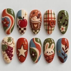 May include: A set of ten almond-shaped press-on nails featuring autumn-themed designs including a red apple, a hot cocoa mug, a plaid pattern with a button, a white rabbit, grapes, stars, flowers, and swirling patterns in shades of red, green, and blue.