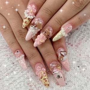 May include: A set of long, almond-shaped acrylic nails with various 3D embellishments. Designs include pink flowers, teddy bears, seashells, and polka dots. Some nails feature a yellow and brown leopard print pattern, while others are a solid pink.