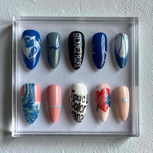 May include: A collection of ten false nails presented in a clear case. The nails showcase diverse designs, such as blue and white marbled effects, a grey nail with a blue line, a black nail with the word "BLUEPRINT", and nails with floral and text designs.