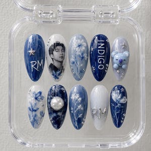 May include: A set of ten almond-shaped press-on nails in a clear plastic case. The designs feature shades of blue and white, including a portrait of a person, the text RM and INDIGO, floral patterns, a 3D koala figure, a pearl, and silver stars.