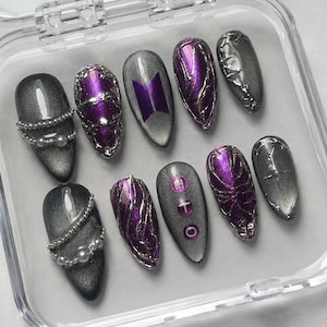 May include: A set of ten artificial nails in a clear plastic case. The nails feature a variety of designs, including silver glitter, purple accents, and a purple logo. Some nails have pearl accents. The nails are oval-shaped.
