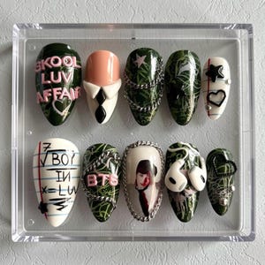May include: A collection of ten artificial nails displayed in a clear case. The nails showcase diverse designs, such as text, chains, stars, hearts, and a portrait. The colour palette includes green, white, pink, and black. Some nails have a matte finish, others are glossy.