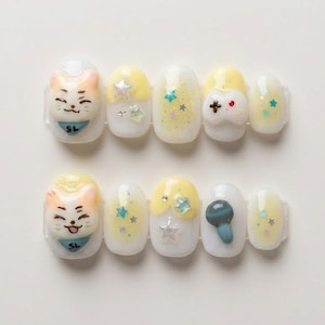 May include: A set of ten press-on nails with a white base, featuring various designs. Some nails have a cat face, others have yellow accents, stars, and a game controller. The nails are decorated with glitter and 3D embellishments.