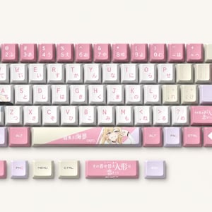 Op de afbeelding: Een mechanische toetsenbord-keycapset met een roze, wit en lichtpaars kleurenschema. De toetsen tonen Engelse letters, Japanse hiragana-tekens en illustraties van anime-personages. Verschillende toetsen bevatten gedrukte Japanse tekst.
