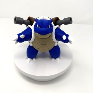 May include: A blue and tan Blastoise figurine with white accents and gray cannons. The figure is posed with arms outstretched, showcasing its iconic design. The figurine is a collectible item.