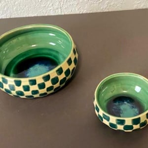 May include: Two ceramic bowls with a green and blue interior and a checkered pattern on the exterior. The larger bowl is on the left, and the smaller bowl is on the right. The bowls are on a brown surface.