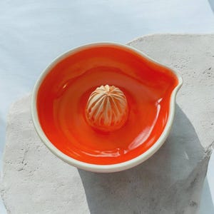 May include: A ceramic citrus juicer with a bright orange interior and a white rim. The juicer features a textured reamer in the center for extracting juice. The design includes a spout for easy pouring.