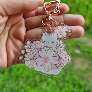 Un pequeño gato con llavero de acrílico con flor de sakura y una pequeña flor de sakura acrílica.