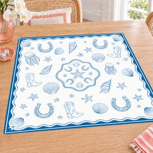 Könnte beinhalten: Eine quadratische weiße Stoffserviette mit einem blauen Druckmuster aus Muscheln, Seesternen, Hufeisen und Cowboystiefeln. Das Design umfasst ein zentrales kreisförmiges Motiv, das von einem gewellten blauen Rand umrahmt wird.