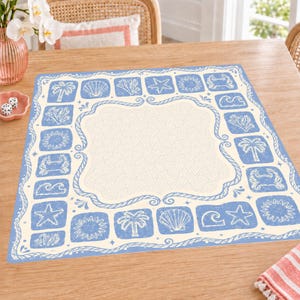 Könnte beinhalten: Ein quadratisches Platzset mit einer cremefarbenen Mitte mit geometrischem Muster, umgeben von einem blauen Rand mit wiederkehrenden weißen Illustrationen von Palmen, Muscheln, Korallen, Seesternen, Sonnen, Krabben und Meereswellen.