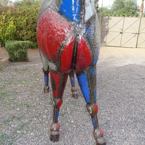 May include: A large metal sculpture of a horse, painted in shades of red, blue, and gray. The horse's legs are bent and the sculpture is standing on a gravel surface.