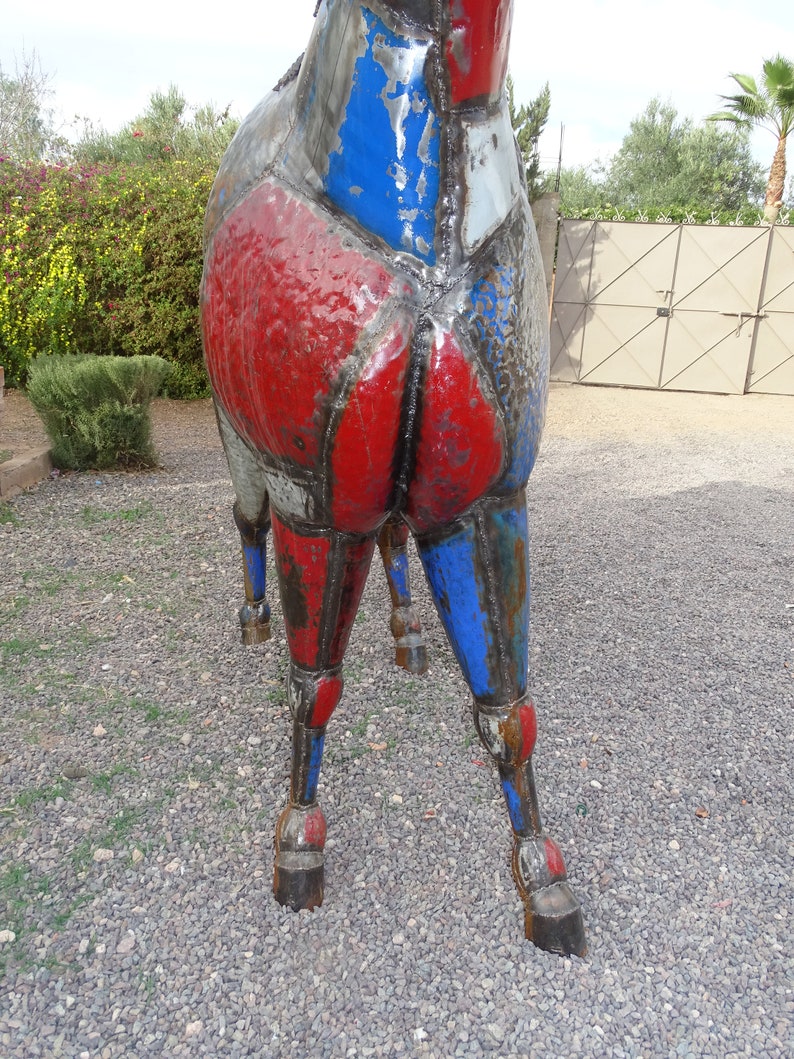 May include: A large metal sculpture of a horse, painted in shades of red, blue, and gray. The horse's legs are bent and the sculpture is standing on a gravel surface.