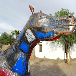 May include: A large metal sculpture of a horse, painted in shades of red, blue, and gray. The horse's head is turned to the side, and its eyes are closed. The sculpture is made of recycled materials and has a rustic, industrial look.