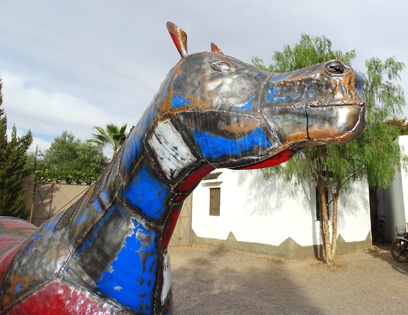 May include: A large metal sculpture of a horse, painted in shades of red, blue, and gray. The horse's head is turned to the side, and its eyes are closed. The sculpture is made of recycled materials and has a rustic, industrial look.