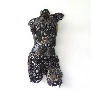 May include: A metal sculpture of a human torso, constructed from various mechanical parts. The sculpture is primarily dark gray and black, with a textured surface created by the arrangement of gears, chains, and other components. The artwork is mounted on a white wall.