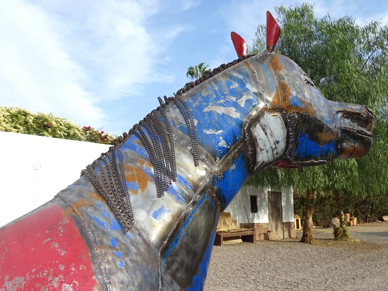 May include: A blue, red, and silver metal sculpture of a horse's head. The sculpture is made of various metal pieces welded together. The horse's mane is made of metal chains.