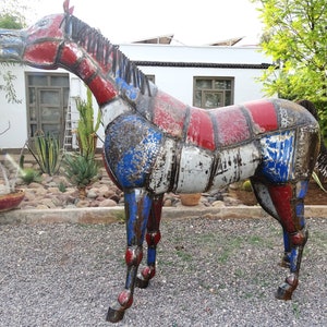 May include: A large metal sculpture of a horse, made from recycled materials. The horse is painted in shades of red, white, and blue, with a distressed finish. The sculpture is standing on a gravel surface.