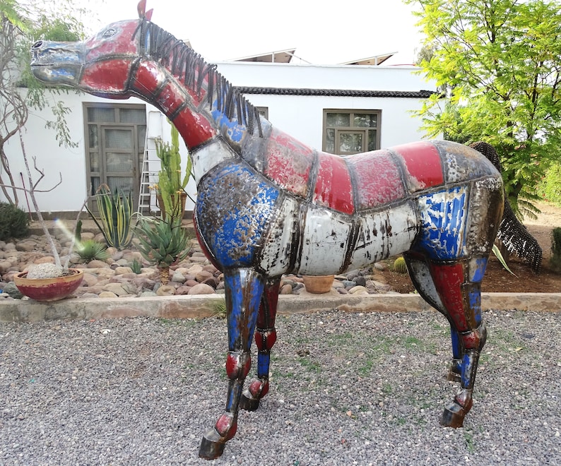 May include: A large metal sculpture of a horse, made from recycled materials. The horse is painted in shades of red, white, and blue, with a distressed finish. The sculpture is standing on a gravel surface.