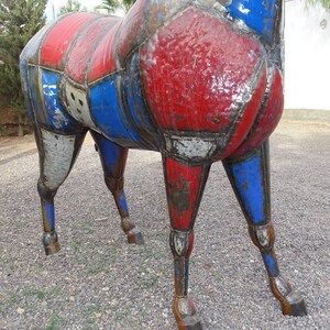 May include: A large metal sculpture of a horse, painted in red, white, and blue. The horse is made of various metal pieces that have been welded together. The sculpture is standing on a gravel surface.