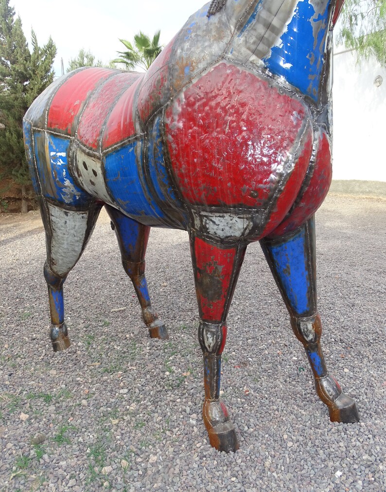 May include: A large metal sculpture of a horse, painted in red, white, and blue. The horse is made of various metal pieces that have been welded together. The sculpture is standing on a gravel surface.