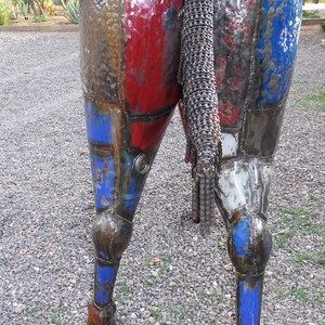 May include: A metal sculpture of a horse's rear end and legs. The sculpture is made of recycled metal and painted in shades of red, blue, and white. The horse's tail is made of a chain.