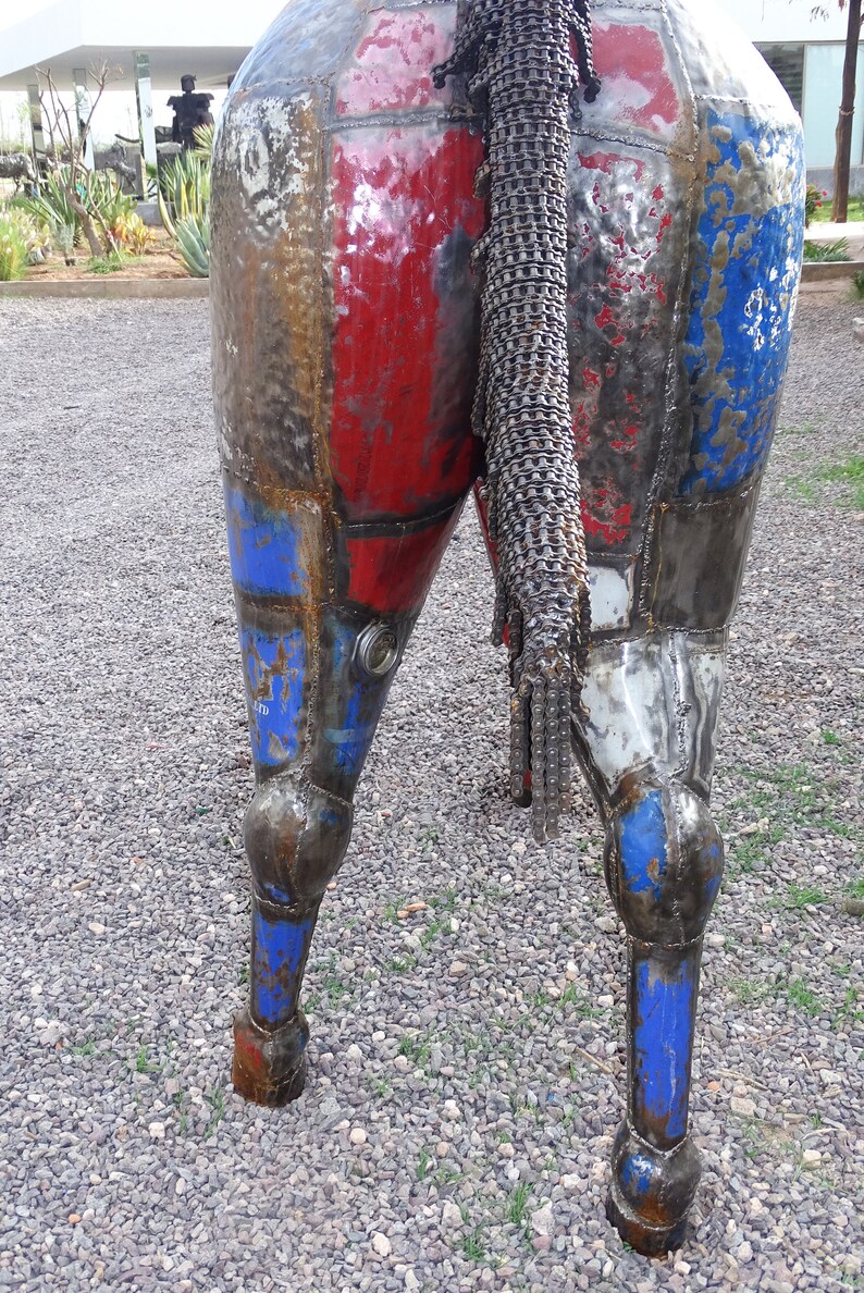 May include: A metal sculpture of a horse's rear end and legs. The sculpture is made of recycled metal and painted in shades of red, blue, and white. The horse's tail is made of a chain.