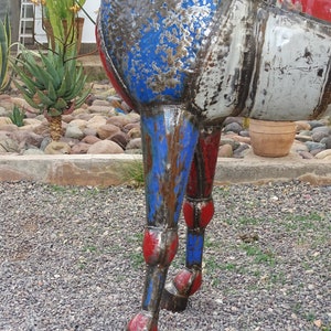 May include: Close-up of a metal sculpture of a horse's legs. The legs are painted red, white, and blue, and have a distressed, weathered look. The sculpture is standing on a gravel surface.