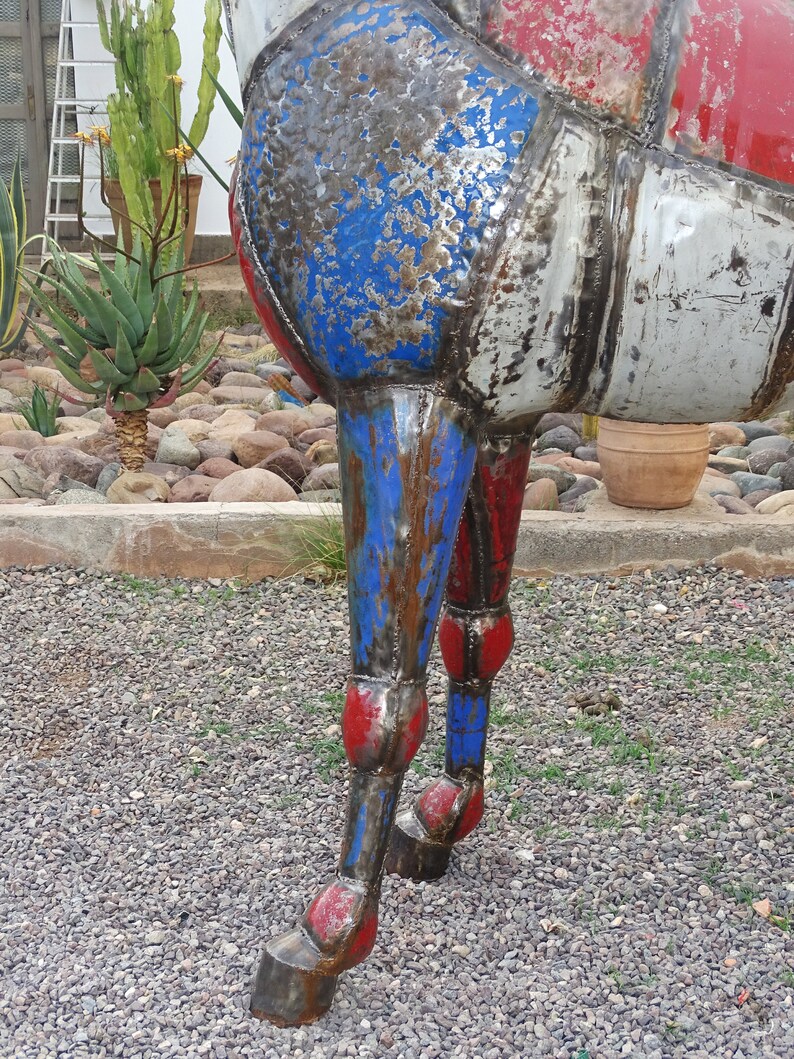 May include: Close-up of a metal sculpture of a horse's legs. The legs are painted red, white, and blue, and have a distressed, weathered look. The sculpture is standing on a gravel surface.
